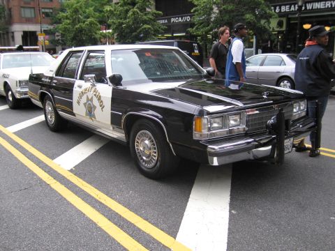 1991 Ford LTD Crown Victoria - black-and-white Highway Patrol sedan with a front push bar parks on a city street as several people stand alongside it near storefronts.