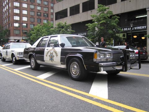 1991 Ford LTD Crown Victoria - black-and-white Highway Patrol sedan with a gold star and front push bar parks along a city street near a winery storefront, another patrol…