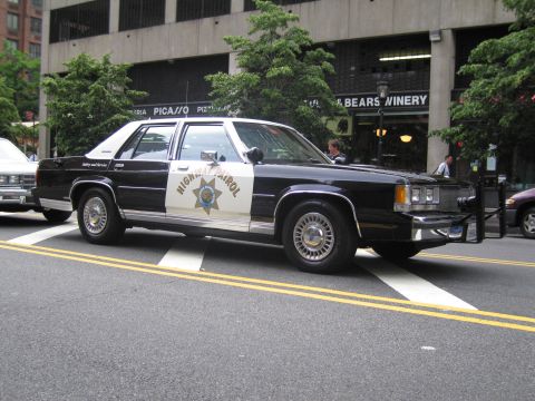 1991 Ford LTD Crown Victoria - black-and-white Highway Patrol sedan with a gold star door emblem parks on a city street, with another patrol car visible behind.
