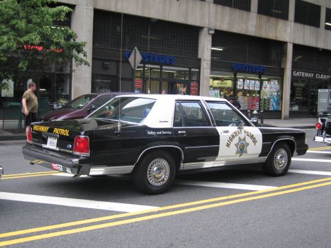 1991 Ford LTD Crown Victoria - black-and-white Highway Patrol sedan with a rear push bar and Safety and Service lettering sits on a city street before a Gristedes store.