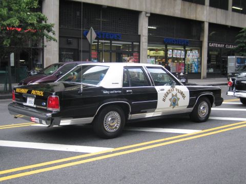 1991 Ford LTD Crown Victoria - black-and-white Highway Patrol sedan with a gold star and Safety and Service lettering is parked along a city curb near storefronts.