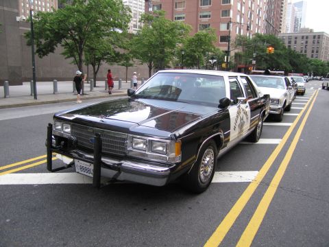 1991 Ford LTD Crown Victoria - black-and-white Highway Patrol sedan with a front push bar parks on a city street as pedestrians walk on the sidewalk behind it.