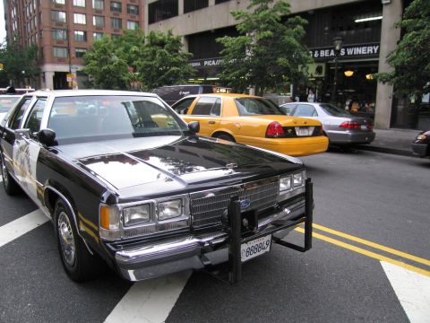 1991 Ford LTD Crown Victoria - black-and-white Highway Patrol sedan with a front push bar sits at a city crosswalk while yellow taxis pass on the street behind.