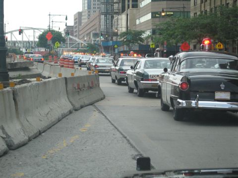1991 Ford LTD Crown Victoria - line of classic police cars, including a black-and-white sedan with tailfins, sits in traffic beside concrete barriers on a city street.