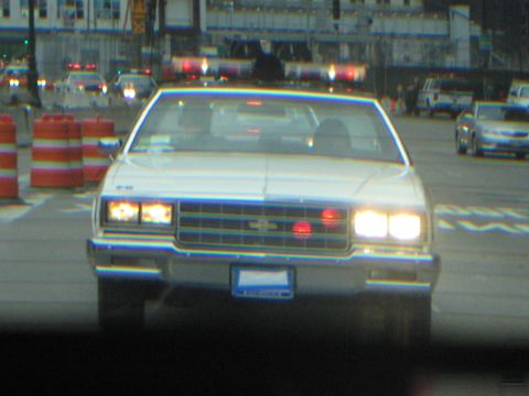 1991 Ford LTD Crown Victoria - white classic sedan with headlights on drives toward the camera on a city street at dusk, with orange barrels and other vehicles nearby.