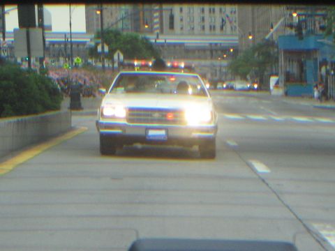 1991 Ford LTD Crown Victoria - white classic car with its headlights on drives down a wide city street at dusk, with flags and buildings lining the route behind.