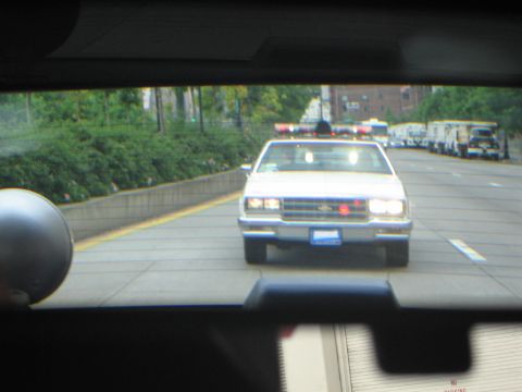 1991 Ford LTD Crown Victoria - View through a car windshield of a white police cruiser with roof lightbar driving toward the camera on a multi-lane highway.
