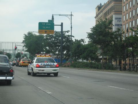 1991 Ford LTD Crown Victoria - white police cruiser with roof lights leads traffic down a broad city avenue beneath a green 10 Ave highway exit sign.
