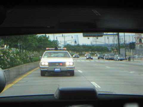1991 Ford LTD Crown Victoria - Through a windshield, a white police cruiser with headlights on drives toward the camera on a multi-lane highway.