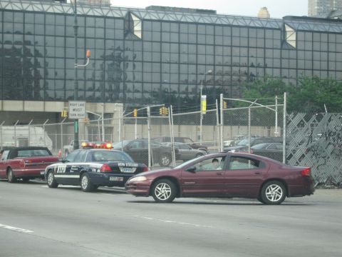 1991 Ford LTD Crown Victoria - black-and-white police car with red roof lights and a maroon sedan are stopped on a city street beside a chain-link fence.