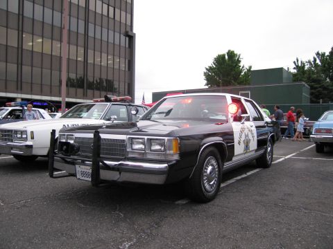 1991 Ford LTD Crown Victoria - black-and-white Ford police sedan with chrome grille, push bar and lit red roof light parked at an outdoor car show.