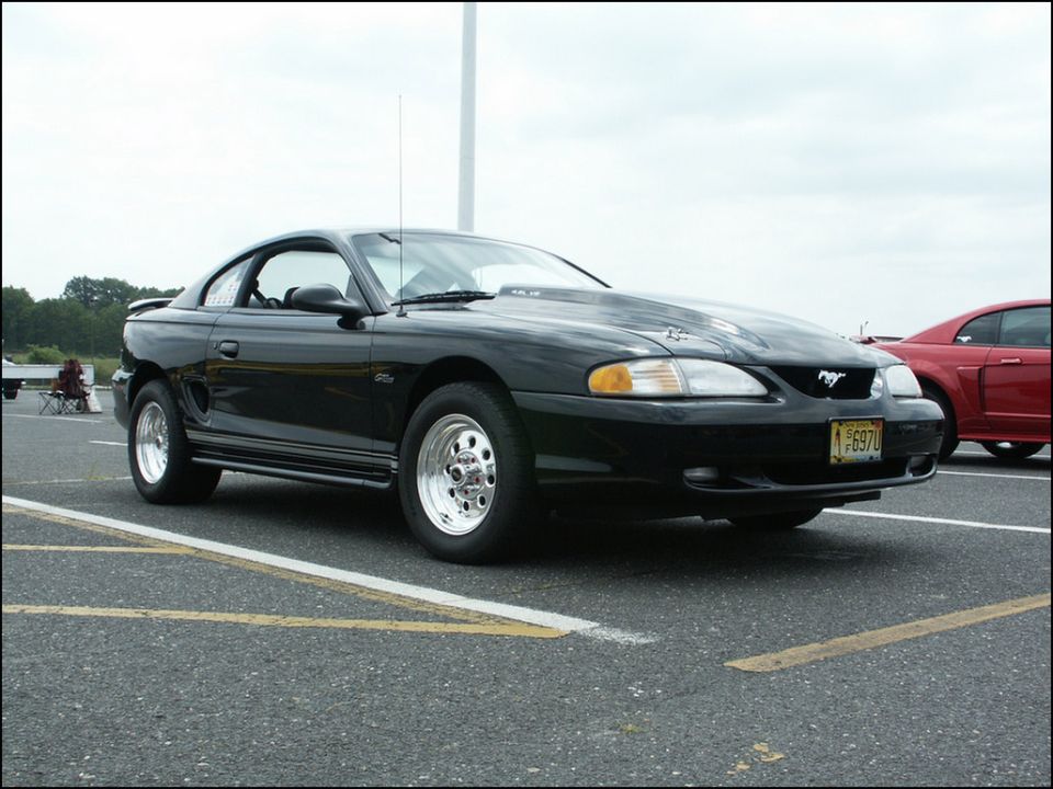 Black Ford Mustang with aftermarket hood and drag wheels. Another red car is parked nearby