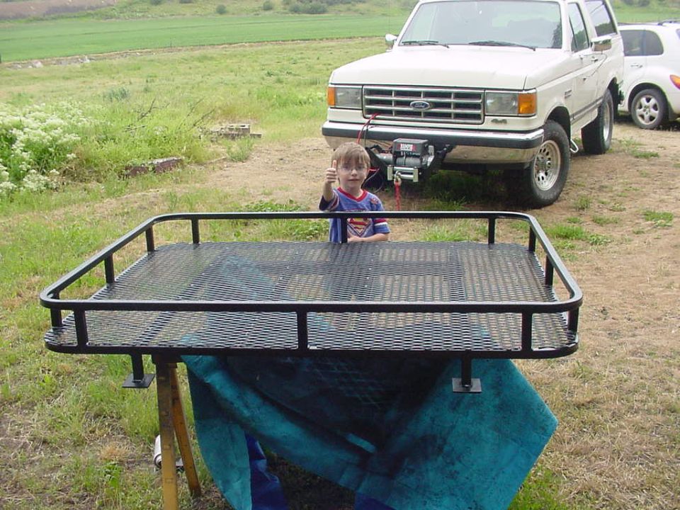 White Ford Bronco SUV with winch behind a black metal cargo rack and a child giving thumbs-up.
