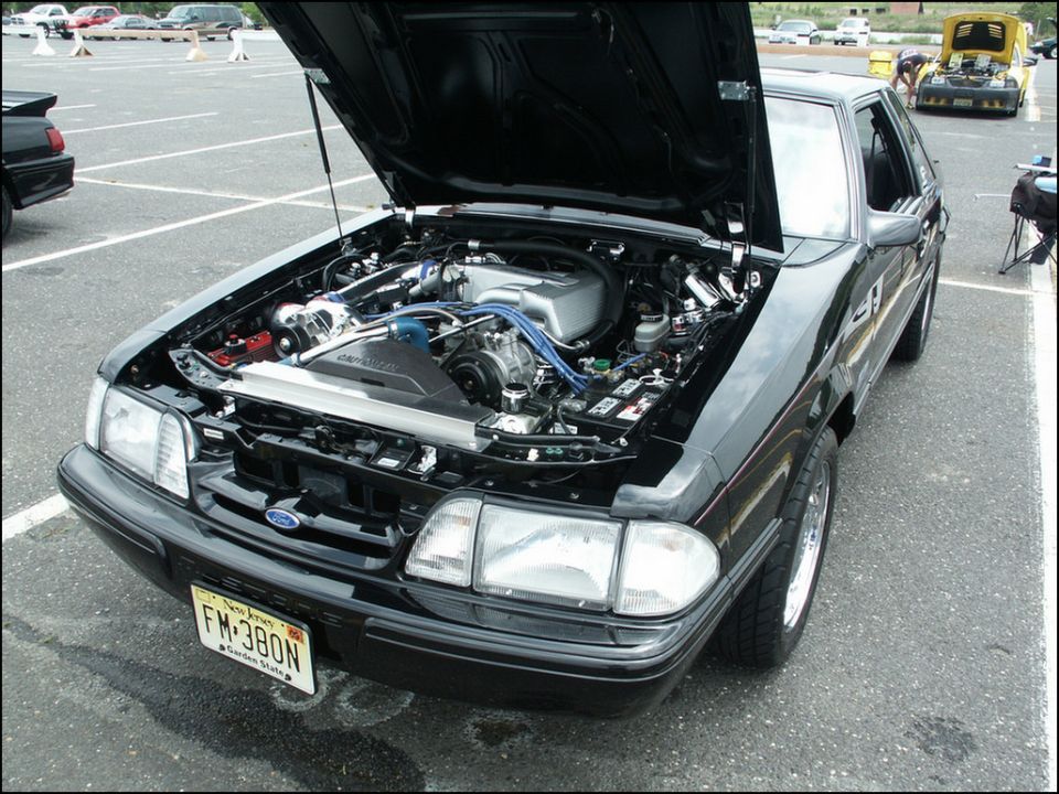Black Ford Mustang with hood open, showing a modified engine. Front view, license plate FM-380N