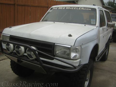 1990 Ford Bronco - white lifted Ford SUV with a tube front bumper, light pods, and TheWebWheeler.com windshield banner sits by a fence.