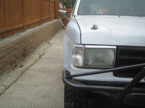 1990 Ford Bronco - Close-up of a white off-road truck's front corner showing a tubular steel bumper, rectangular headlight, and mud-terrain tire near a wood f…