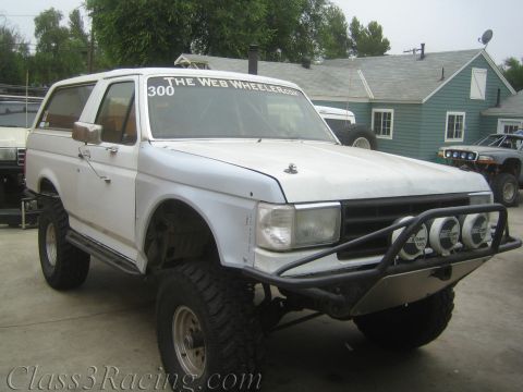 1990 Ford Bronco - Lifted white two-door Ford Bronco on large mud tires with a tubular front bumper and a windshield banner reading The Web Wheeler.