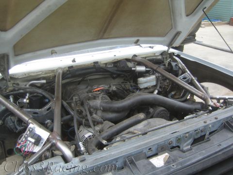 1990 Ford Bronco - Engine bay under a raised hood showing a V8 engine, radiator hoses, alternator, and tubular cage framing around the compartment.