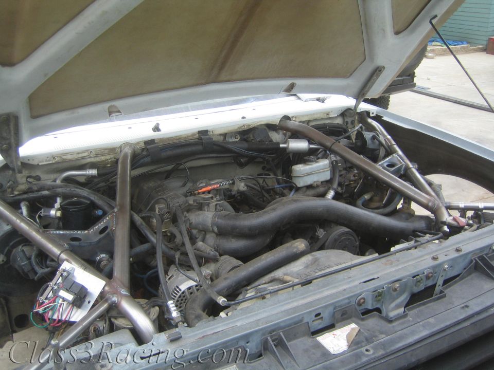 Open engine bay of a vehicle with V8 engine, hoses, and tubular roll cage framing