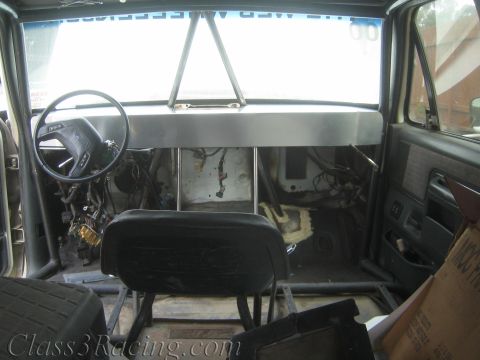 1990 Ford Bronco - Stripped truck cab showing a steering wheel, fabricated aluminum dash panel, a single black bucket seat, and tubular cage tubes.