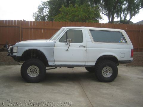 1990 Ford Bronco - Side profile of a lifted white two-door SUV with a hardtop on large mud tires, parked on concrete beside a wood fence.
