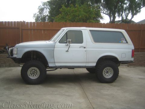 1990 Ford Bronco - Side view of a lifted white two-door SUV with a removable hardtop and large mud tires parked on a concrete driveway by a fence.