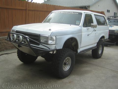 1990 Ford Bronco - Front three-quarter view of a lifted white two-door SUV with a tubular bumper, three round driving lights, and mud tires.