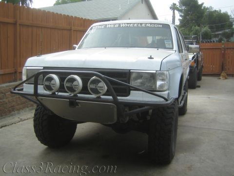 1990 Ford Bronco - Front view of a primer-gray off-road truck with a tubular bumper, three round Pro Comp lights, and a windshield banner reading The Web Whee…