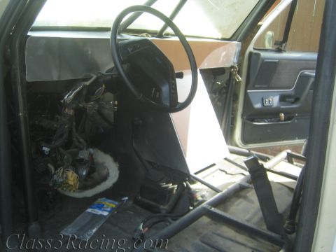 1990 Ford Bronco - Stripped truck cab interior showing a steering wheel, fabricated sheet-metal dash, exposed wiring, tube cage, and a gray door panel.