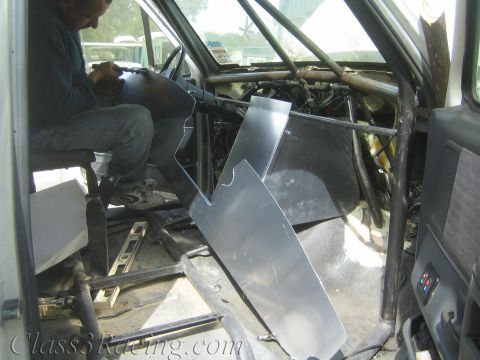 1990 Ford Bronco - person crouches inside a gutted vehicle cab fitting bare sheet-metal panels among the tube cage and exposed components.