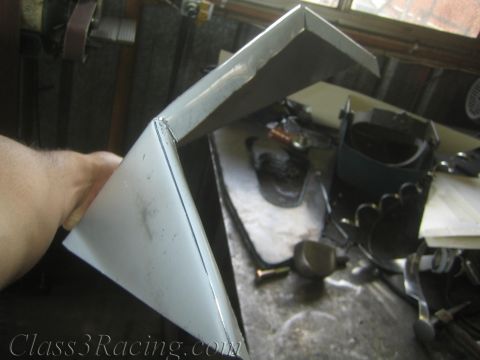 1990 Ford Bronco - hand holds up a folded and welded bare sheet-metal bracket in a workshop with scattered tools and parts behind it.