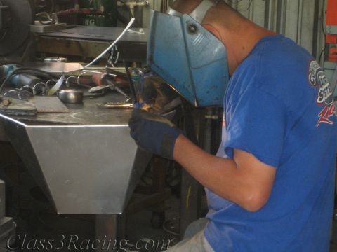 1990 Ford Bronco - person in a blue shirt and blue welding helmet TIG-welds a bare sheet-metal fabricated part at a workshop bench.