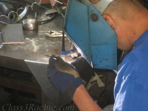 1990 Ford Bronco - welder in a blue shirt and blue helmet welds small metal pieces clamped at a steel workbench, viewed over the shoulder.