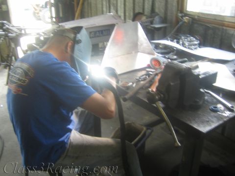 1990 Ford Bronco - seated welder in a blue shirt and helmet welds a part held in a bench vise, the arc casting bright light in a cluttered shop.