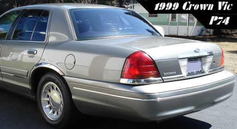 1999 Ford Crown Victoria - Three-quarter rear view of a light gold/champagne Ford Crown Victoria sedan parked on a driveway beside a mobile home, showing the full-wid…