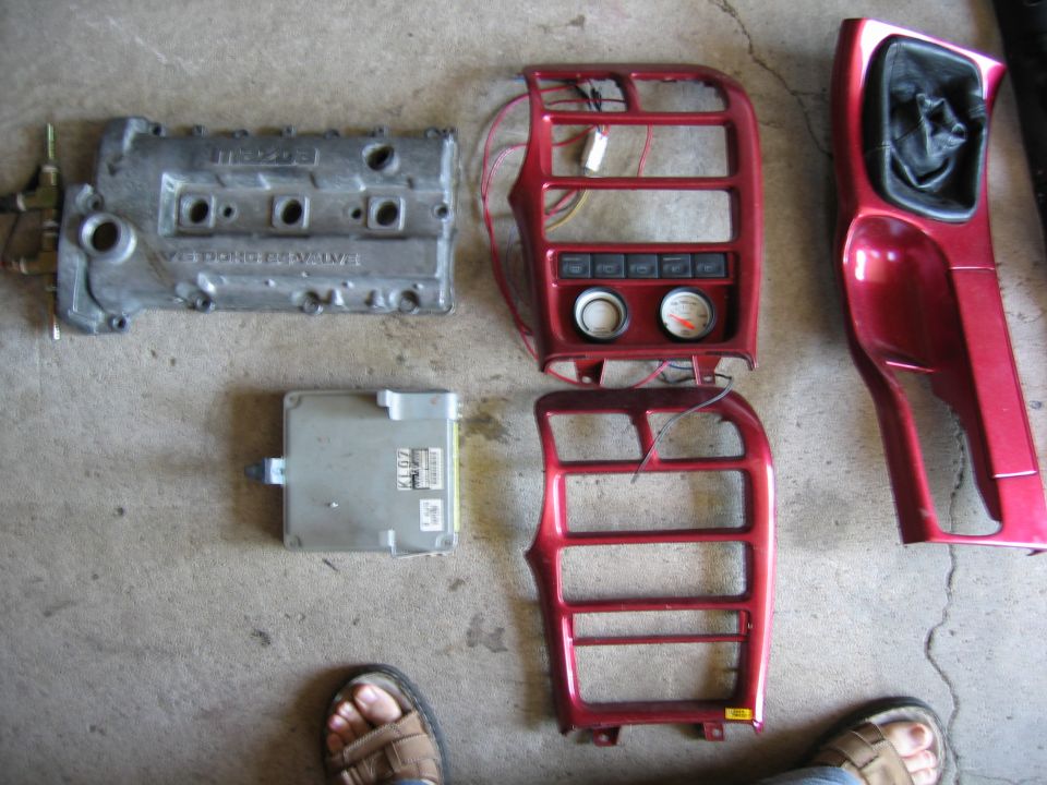 Mazda engine valve cover, ECU, and red interior trim pieces laid out on concrete