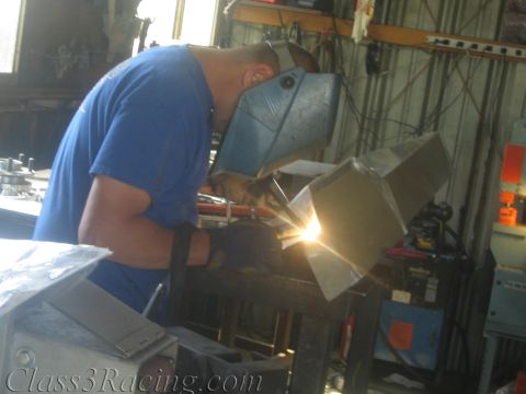 1990 Ford Bronco - welder in a flip-up helmet and blue shirt fuses aluminum pieces on a workbench in a fabrication shop.