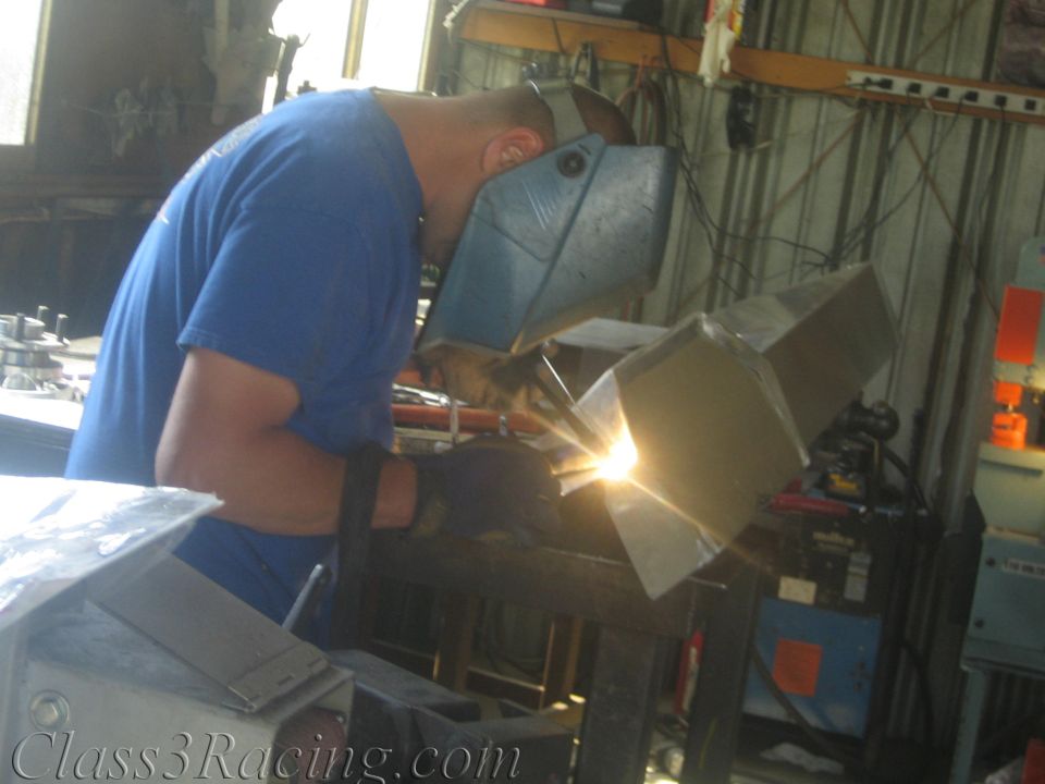 Welder wearing a helmet joining aluminum parts at a shop bench