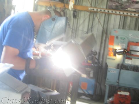 1990 Ford Bronco - welder in a blue shirt and helmet works an aluminum assembly, the arc producing an intense burst of light.