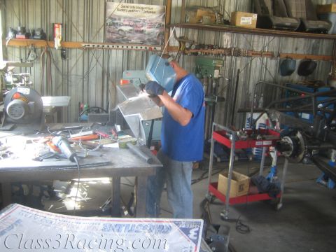 1990 Ford Bronco - welder works at a bench in a cluttered shop, with a chassis on a lift and a banner overhead.