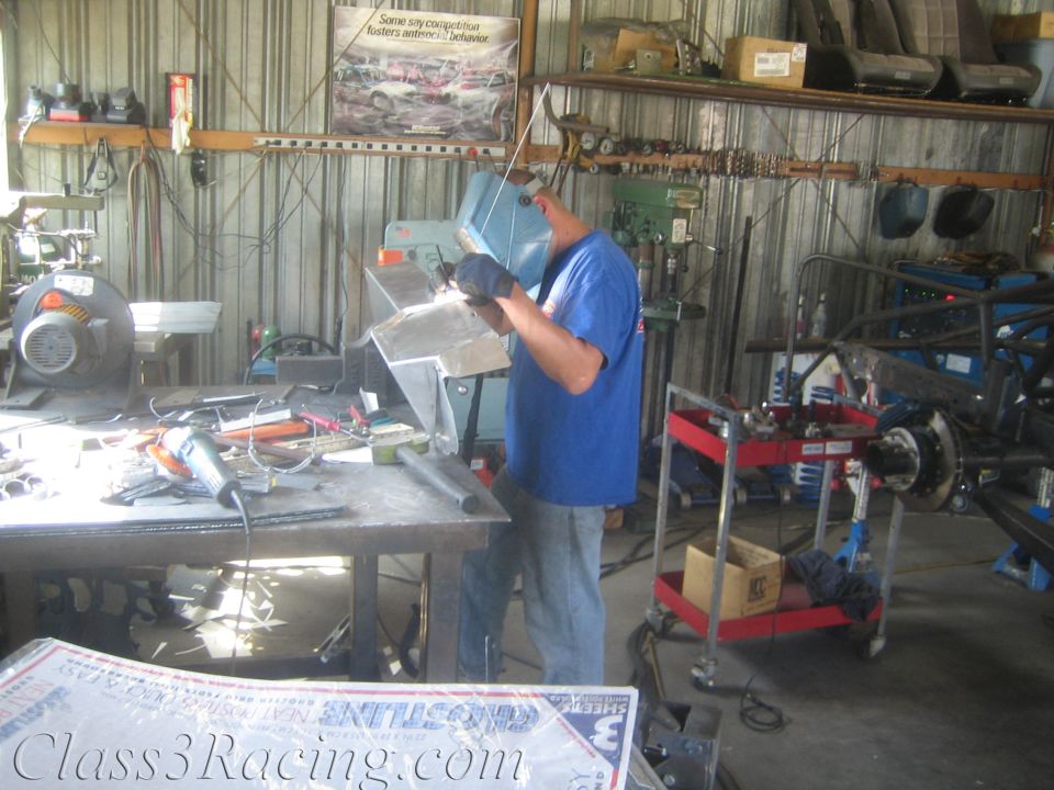 Welder at a bench in a busy fabrication shop with vehicle parts