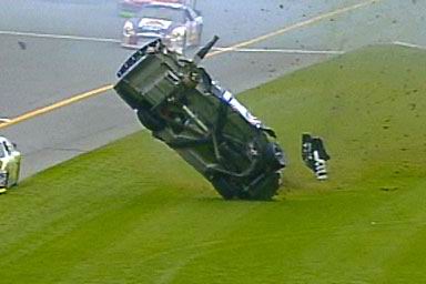 1981 Ford Bronco - A stock car has crashed and is on its side on the grass next to the track.