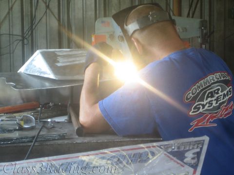 1990 Ford Bronco - welder in a blue racing-logo shirt fuses an aluminum panel, the arc flaring brightly at the bench.