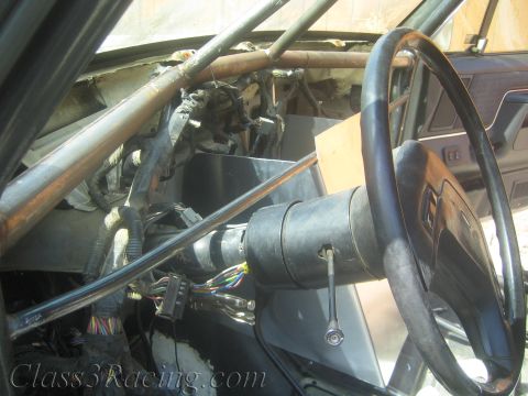 1990 Ford Bronco - Exposed steering column and a wood-rimmed steering wheel beside roll-cage tubing in a gutted vehicle interior.