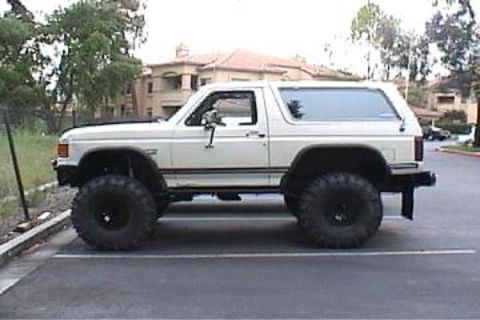 1991 Ford Bronco - This modified SUV is parked in a lot with a building and trees in the background.