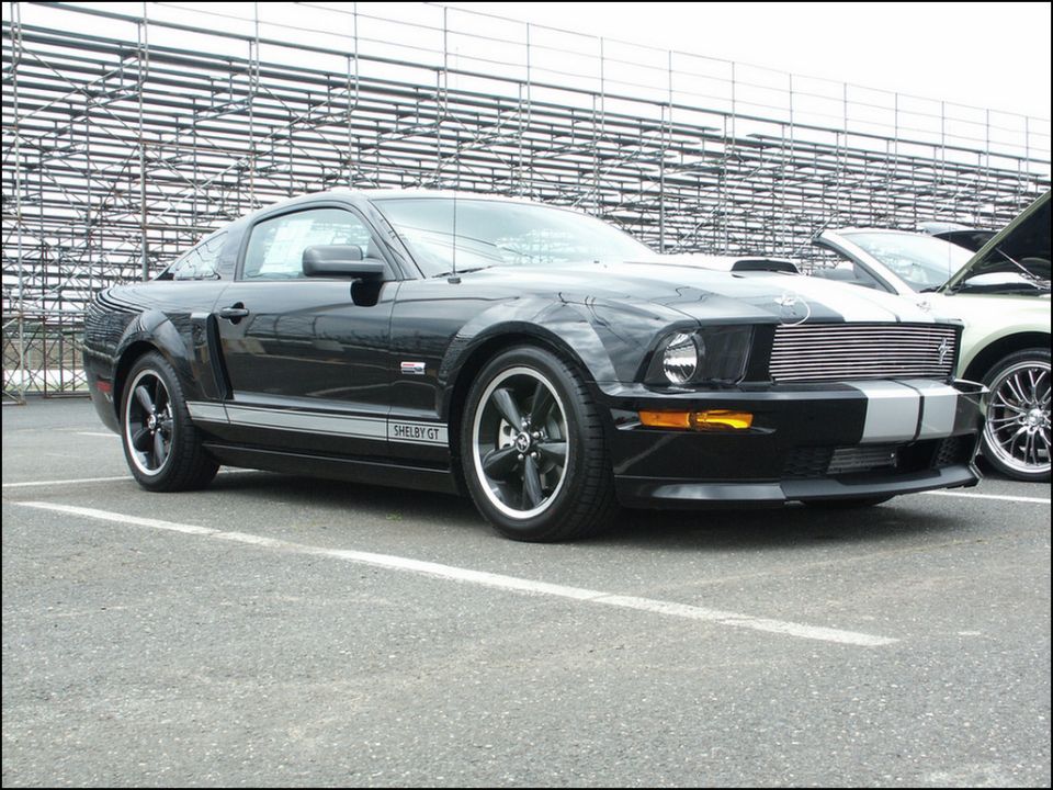 Black Ford Mustang Shelby GT with silver racing stripes parked in front of stadium bleachers. Another car is visible to the right