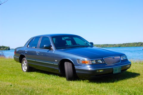 1985 Mercury Grand Marquis - A blue sedan sits on a grassy area next to a body of water under a clear blue sky.