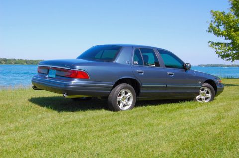 1985 Mercury Grand Marquis - A blue sedan is parked on a grassy area next to a body of water under a clear blue sky.