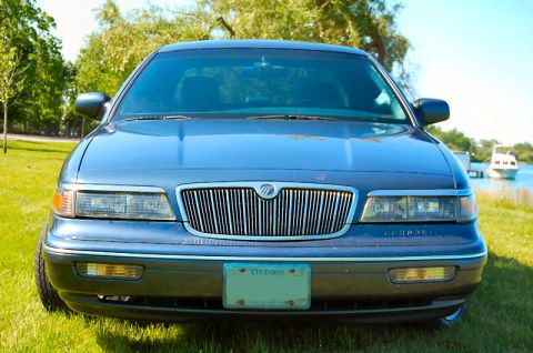 1985 Mercury Grand Marquis - A blue sedan is parked on a grassy area near a body of water with a boat in the background.