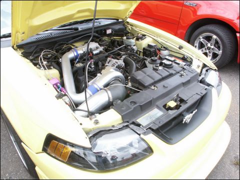 2001 Mercury Grand Marquis - Engine bay of a yellow car a turbocharger and custom intake piping, with a red truck in the background.